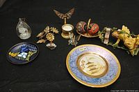 Lot overview showing the enamel plate, Swarovski crystal bouquet, butterfly brooch, various small figurines and decorative trinkets arranged on a dark surface.
