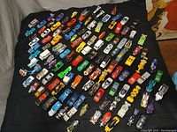 Wide view of over 120 assorted Hot Wheels vehicles laid out on a black surface showing a variety of colors and models including cars, trucks, motorcycles, and construction vehicles.