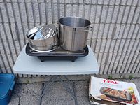 Photo of T-Fal electric grill, large stainless steel stock pot and three metal bakeware pans on small table outdoors, includes grill box on ground.
