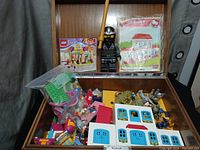 Wooden box open showing assorted LEGO sets, loose bricks in bags, LEGO Star Wars and other minifigures, and instruction manuals for LEGO Friends and Hello Kitty Mega Bloks sets.