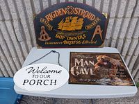 Lot of three decorative signs displayed on white table outdoors against a stone wall: the double-sided nautical RIGDEN & STROUD wooden sign, white oval Welcome to Our Porch sign, and rectangular Man Cave sign with bear graphic.