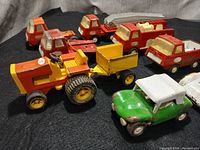 Photo showing nine vintage pressed steel Tonka trucks and toy vehicles arranged on a black surface including a yellow tractor with trailer, red fire truck, green car, white race car, and various small trucks.
