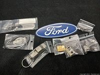 Photo showing 9 Ford branded metal keychains and pins sealed in plastic bags, including commemorative items for 50 and 100 years and original tagged keychain.