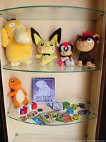 Main lot showing four larger plush toys displayed on a glass shelf: Psyduck, Pichu, red-faced hedgehog, and brown monkey plush.