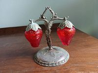 Front view showing the two red strawberry salt and pepper shakers on the ornate metal stand with leaf designs and aged patina.