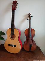 Photo showing small guitar with natural wood top and red sides standing next to varnished brown violin.