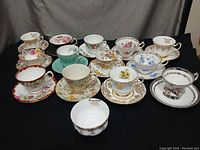 Photo of 13 assorted tea cups and saucers arranged on black fabric, showing floral and gold trimmed designs.