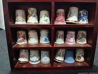Wooden display shelf holding 18 ceramic decorative thimbles in four compartments, showing various floral and scenic designs on the thimbles