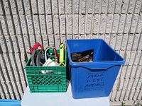 Green milk crate filled with assorted hand tools and a blue plastic bin beside it on a white table against a block wall.