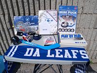 Photo showing the complete lot of Toronto Maple Leafs hockey collectibles including scarf, mini puck collection, keychain, stat book, helmet souvenir, bootie, and other small items placed outdoors on a folding table.