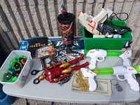 Full view of toy lot displaying Iron Man action figure, Beyblades in a white tub, toy guns, Spider-Man cup, airplane toy, toy scissors, and various toy accessories on a table outdoors with a block wall background.