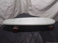 Side profile photo of the white skateboard deck with red wheels on a dark cloth background, showing condition and shape.