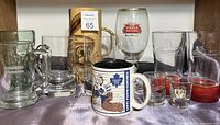 Group of assorted glasses and mugs displayed on a surface including shot glasses, beer glasses, wooden stein, and branded items clear and colored