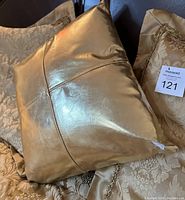 Close-up image of the shiny gold decorative throw pillow showing fabric texture and stitching with floral pattern pillows in the background.