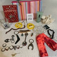 Lot contents showing costumes rings, keychains, bracelets, pins, Clinique scrub creme, cosmetic bag, and Canucks shot glass displayed on countertop