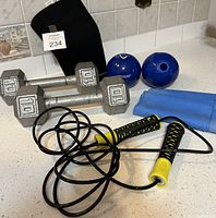 Photo showing two silver metal hex dumbbells each weighing 10 lbs, black and yellow handled skipping rope, two blue exercise balls, blue elastic exercise band, and black knee brace size medium on a white kitchen counter with tiled backsplash.