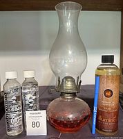 Large vintage oil lamp with rounded red base and clear glass chimney. Accompanied by fuel bottles and a blue pen for scale.