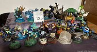 A photo showing an assortment of 20 Skylanders figures with different designs and colors, arranged on a surface with a maxsold auction label in the middle.
