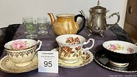 Photo showing the full collection including three teacups with saucers, silver plated teapot, orange ceramic teapot without lid, and three green shot glasses on a dark cloth background.