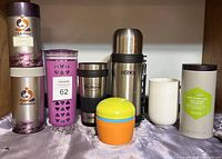 Front view of all items including four Teavana tea containers, a white ceramic mug, thermos, travel cup and colorful plastic cup.
