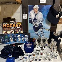 Overview of lot displaying Darcy Tucker painting, visors, mini Stanley Cups, mini mugs, and sticker album on a tiled kitchen counter and nearby surfaces.