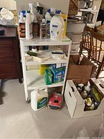 Photo showing white plastic three-tier cleaning cart with multiple cleaning products including sprays, liquids, boxes, and handheld brushes around and on it.