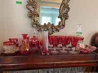 Overview of cranberry glass items including goblets, vases, decanter, bowls, compote, pitchers, arranged on wooden surface beneath ornate mirror