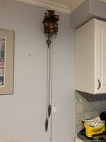 Full view of the cuckoo clock mounted on wall, showing pine cone weights and pendulum chains hanging down, with shadow and wall background.