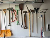 Full view of gardening tools mounted on garage wall showing all items including shovels, rakes, saw, pruners and brush.