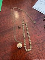 Full view of three gold items on wood surface: rope chain bracelet, fine link chain with round locket, and maple leaf pin