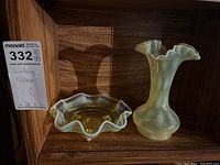 Photo showing two matching yellow iridescent glass pieces: a ruffled footed bowl and a ruffled vase, both on a wooden shelf.