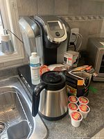 Photo showing Keurig coffee machine with digital display, stainless steel 2.0 carafe, multiple Tim Hortons K-Cup pods, unopened pod boxes, and descaler bottle on kitchen counter next to sink.