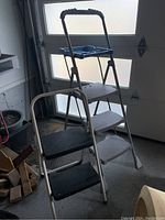 Photo showing the Cosco three-step ladder with blue plastic tool tray in the background and the two-step stool with black treads in front of it inside a garage.