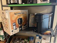 Photo showing the Nespresso Vertuo Next coffee machine on a shelf next to its original box and accessories including a milk frother