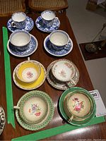 Top view of eight teacups with matching saucers arranged on a wooden table. Variety of colors and floral patterns visible.