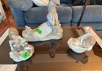 Photo showing all three figurines on a glass table with sofa in background. Included are two Lladro figurines and one larger Porceval figurine.