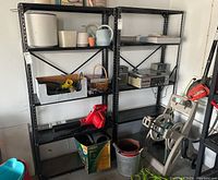 Two black metal five-tier storage shelves loaded with various garage items such as pots, a watering can, a red leaf blower, tools, and containers. Shelves show visible rust and wear.