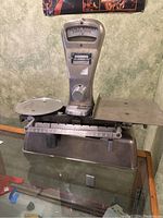Front view of antique metal balance scale showing dial indicator, weighing platforms, and adjustment knobs.
