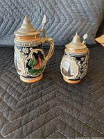 Two decorative ceramic beer steins with pewter lids and scenic relief designs shown from front and angled side.