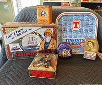 Photo shows seven vintage collectible tins of various brands arranged on a black padded surface. Visible tins include Dominion Royal Bicycle Repair Kit small tin (Canadian made), Fisherman's Friend large tin with fisherman artwork, Smokin Joes Racing tin, Kardomah Orange Spiced Tea tin, and Tennents Lager tray.