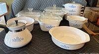 Overview of the entire Corning Ware lot showing multiple white glass-ceramic cookware pieces with blue cornflower pattern, including dishes and lids arranged on a black quilted surface.