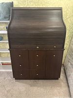 Front view of closed roll-top secretary desk with 6 drawers and one cabinet door, brass knobs, dark wood finish.