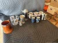 Photo showing 14 assorted collectible mugs and cups on black padded surface including ceramic stein, glass mug, metal tankards, and smaller cups.