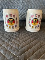 Two white ceramic German style beer stein mugs with German eagle and coat of arms designs side by side.