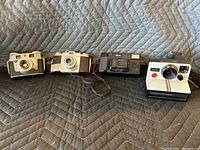 Four vintage cameras on fabric background, showing front and top views, including Kodak and Polaroid models