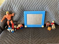 Lot showing all toys arranged on a quilted surface including Etch A Sketch, Playmobil figures, Garfield figure, Nissan forklift, and large wrestler action figure.