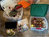 Multiple clear plastic bins containing various Christmas decorations including fabric dolls, small ornaments, and wrapping materials on a concrete floor.
