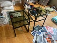 Pair of black metal frame end tables with dark tinted glass tops and lower shelves placed on hardwood floor surrounded by scattered papers and household items.