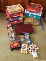 Photo of assorted boxed board and card games stacked on the floor showing titles including Munchkin, Eyecatcher, Jenga, Sequence, Raj, Dominoes, Mental Floss, Mille Bornes, Blink, Quelf, Casio, and The Worst Case Scenario Junior.