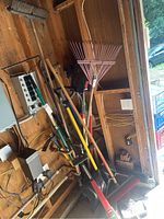 Photo showing various garden hand tools including metal and plastic rakes, shovels, broom, and extension pole leaning against a wooden wall.